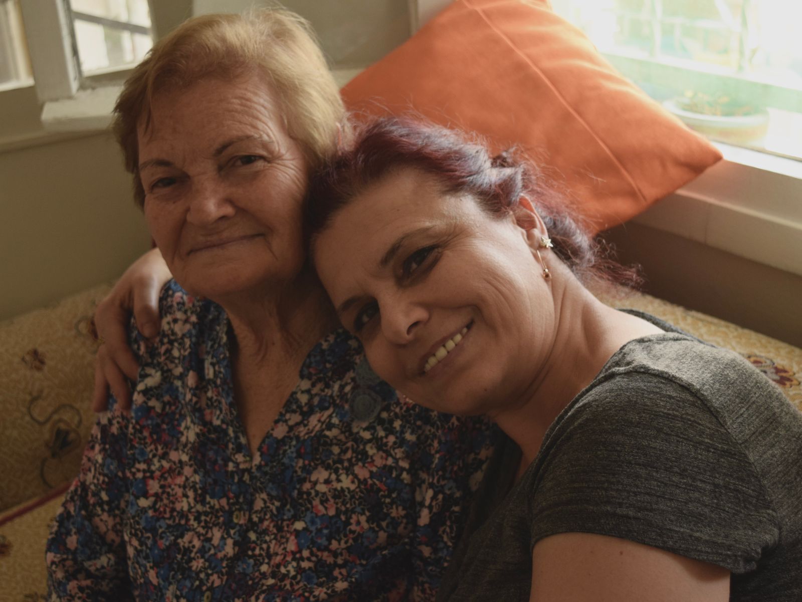 A grown daughter holds her mother close on a sofa at home, both of them smiling