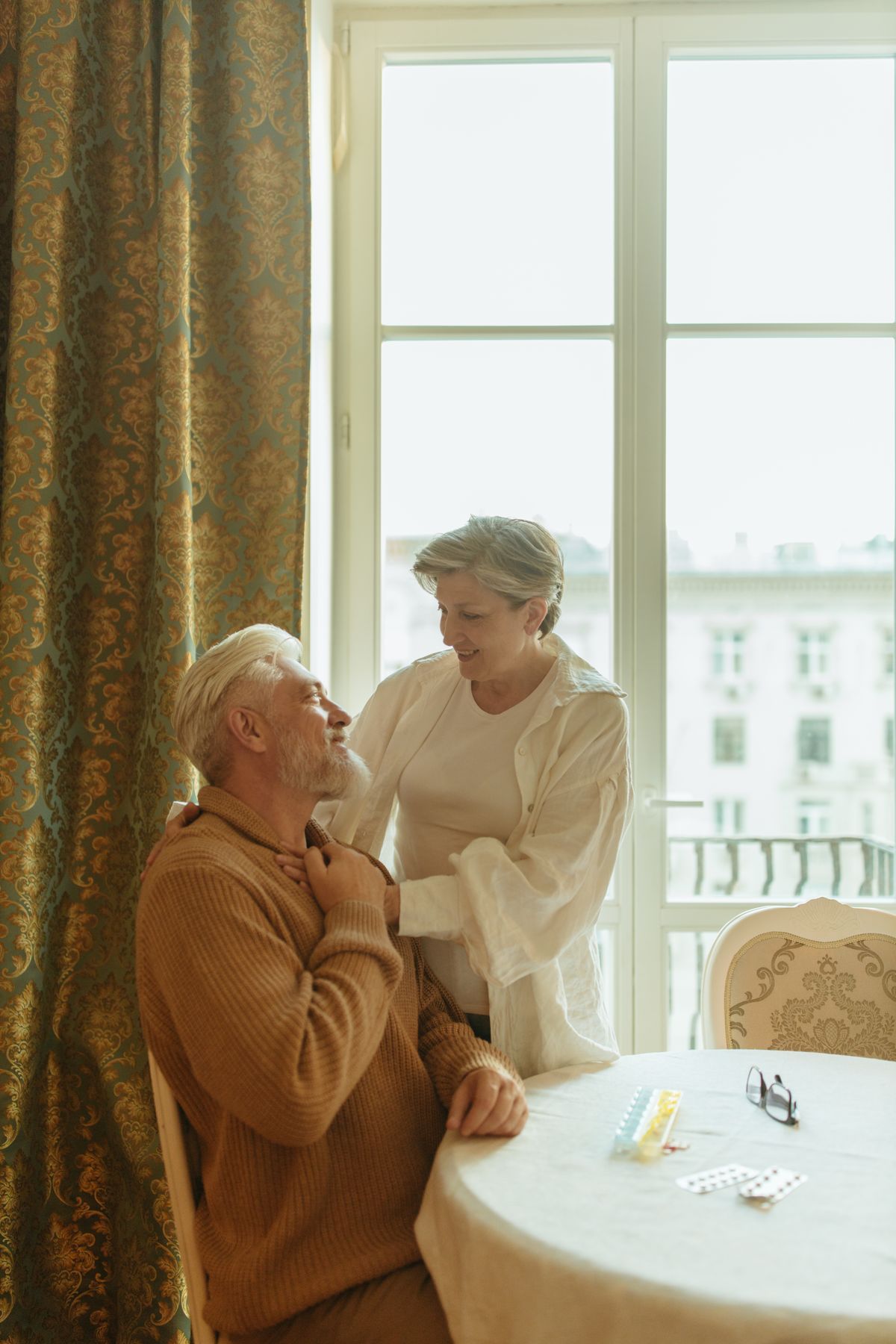 Two people sit and talk quietly beside a tall window in late afternoon light
